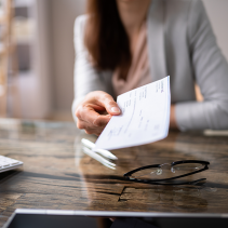 Person handing over a check across a desk with glasses and a keyboard.